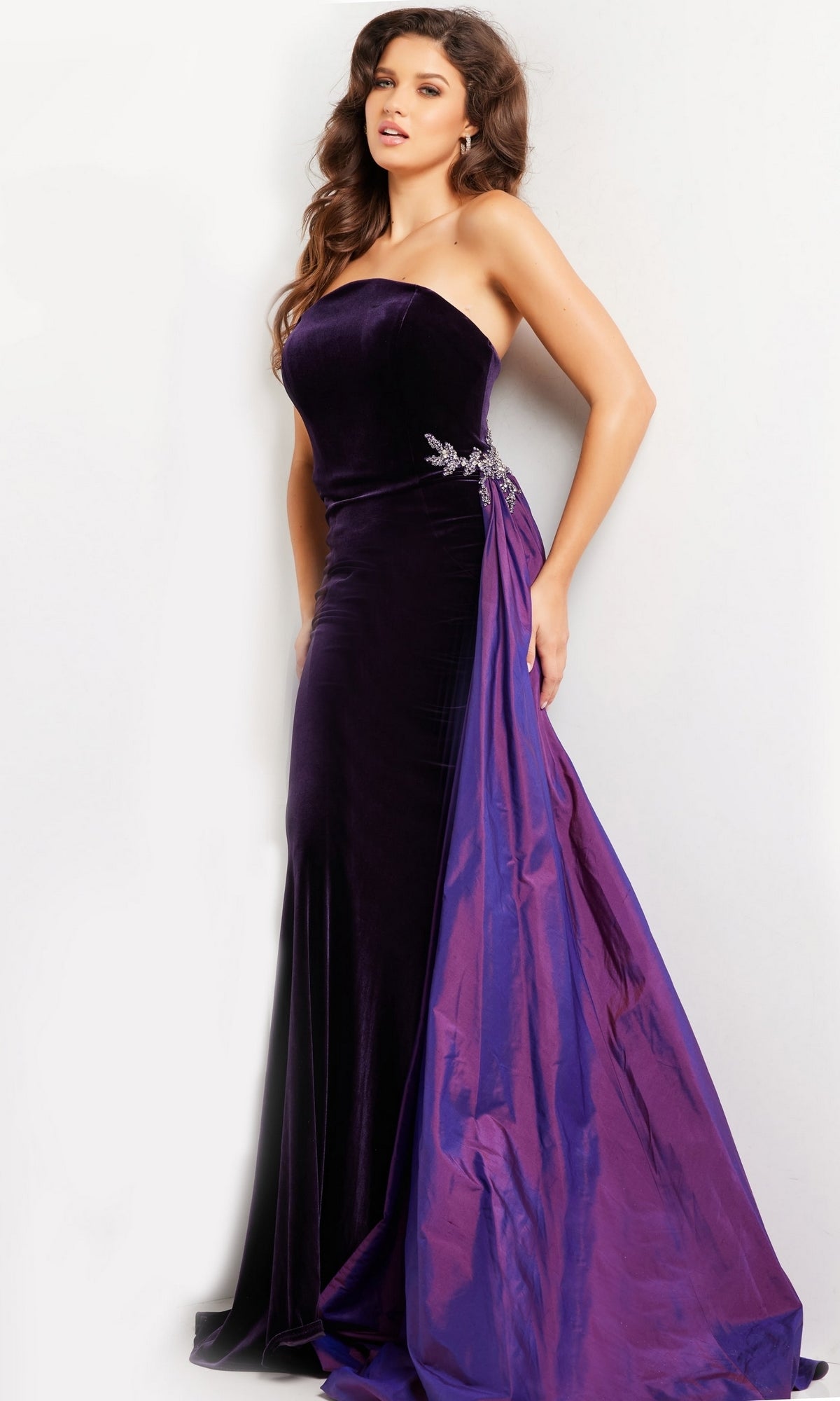 A woman models the Jovani Strapless Long Velvet Prom Dress 26116 in deep purple with embellished detail and shiny overskirt, standing against a plain light background. This elegant formal gown radiates sophistication and timeless style.
